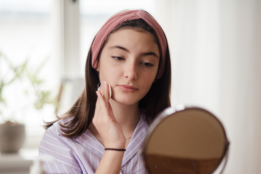 Teen girl with long brown hair wearing a pink headband and striped top, gently applying skincare with a cotton pad while looking in a mirror.